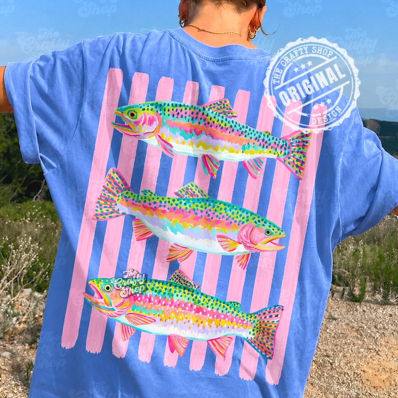 May include: A light blue t-shirt with a design of three vibrant rainbow trout. The fish are set against pink vertical stripes. The shirt has a circular logo that says "The Crafty Shop Original Design".