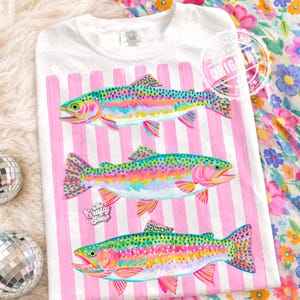 May include: White t-shirt featuring a colourful illustration of three rainbow trout. The fish are painted with vibrant hues of pink, yellow, blue, and green. Vertical pink stripes are in the background. The text "The Crafty Shop" is visible.