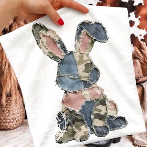 Pode incluir: Camiseta branca com um design de coelho em patchwork. O coelho é feito de vários tecidos em tons de azul, rosa, verde e padrões de camuflagem. O design tem uma borda desgastada.