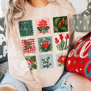 Könnte beinhalten: Ein beiges Langarm-Sweatshirt mit einem Raster aus neun quadratischen Illustrationen von Weihnachtspflanzen und Blumen, darunter Stechpalmen, Weihnachtssterne und rote Beeren, mit einem kreisförmigen Wasserzeichen-Logo unten links.