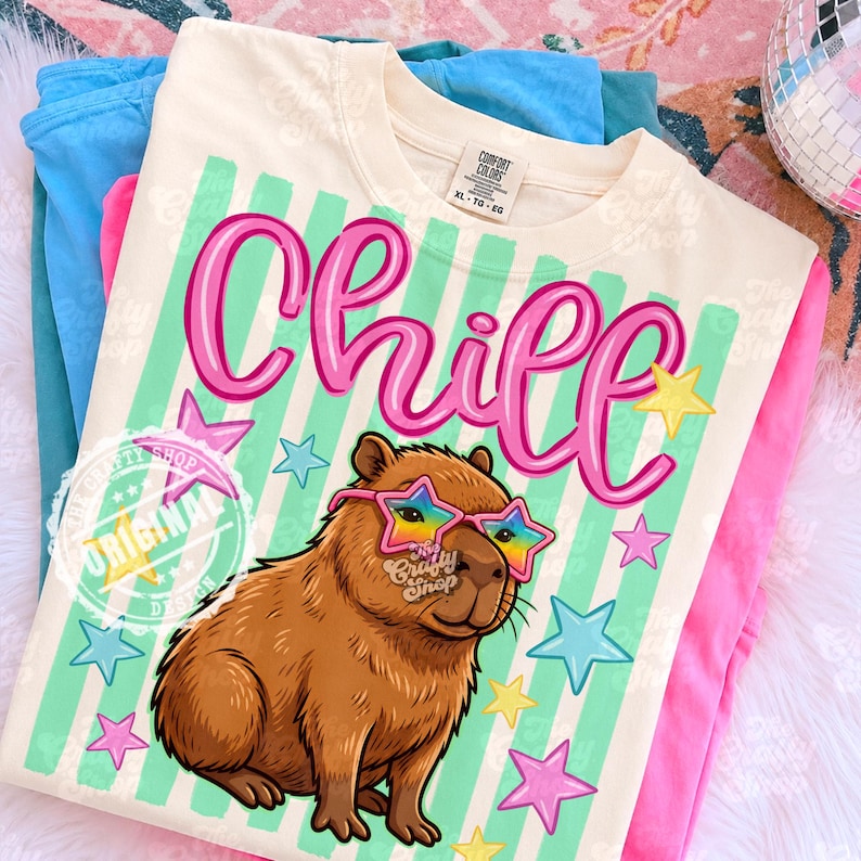 Op de afbeelding: Cr&egrave;mekleurig T-shirt met het woord "Chill" in roze script, een cartoon capibara met een bril in de vorm van een ster en sterren. Het shirt heeft een groen-witte gestreepte achtergrond en de tekst "The Crafty Shop Original Design".