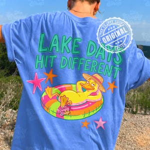 May include: A light blue t-shirt with the text "LAKE DAYS HIT DIFFERENT" in green. The shirt features a cartoon duck wearing a hat and floating on an inner tube. Pink and orange stars are also on the shirt. The shirt has a "The Crafty Shop" logo.