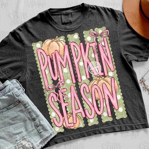May include: A charcoal gray t-shirt featuring a graphic print with the text PUMPKIN SEASON in large pink letters. The design includes three light orange pumpkins, pink bows, and a background of vertical sage green stripes with white polka dots.