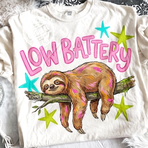 Puede incluir: Camiseta color crema con las palabras "LOW BATTERY" en rosa, con una ilustraci&oacute;n de un perezoso. El perezoso es marr&oacute;n con detalles en rosa y naranja, descansando en una rama. Estrellas turquesas y verde lima rodean el texto y el perezoso.