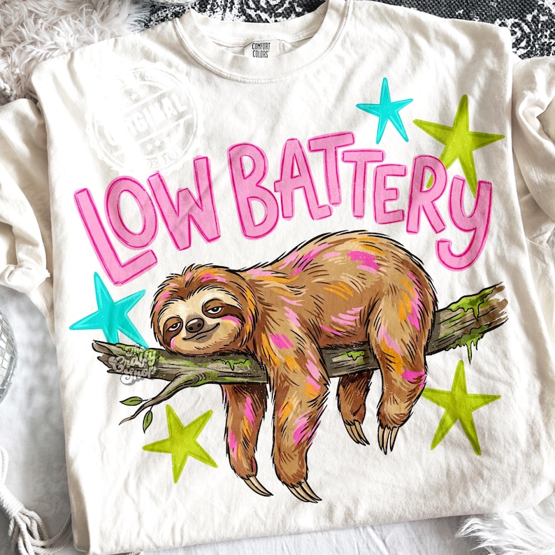 Puede incluir: Camiseta color crema con las palabras "LOW BATTERY" en rosa, con una ilustraci&oacute;n de un perezoso. El perezoso es marr&oacute;n con detalles en rosa y naranja, descansando en una rama. Estrellas turquesas y verde lima rodean el texto y el perezoso.