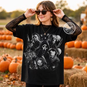 May include: A black t-shirt featuring a white graphic print of various horror movie characters, including a masked figure with a knife, a clown, and a doll. The shirt has long, sheer black sleeves with a spiderweb pattern and a crew neckline.
