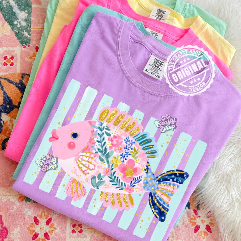 May include: A stack of colorful t-shirts featuring a whimsical fish design. The shirts are in shades of pink, green, yellow, and purple. The fish is decorated with floral patterns and glitter accents. The text "The Crafty Shop" and "ORIGINAL DESIGN" are visible.