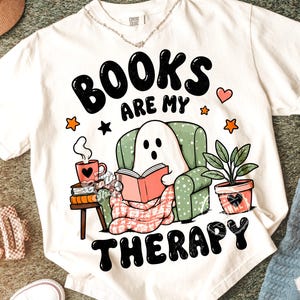 Pode incluir: T-shirt branca com a frase "BOOKS ARE MY THERAPY". O design apresenta um fantasma lendo um livro em uma poltrona verde, uma xícara de café, uma planta em vaso, estrelas e um coração.