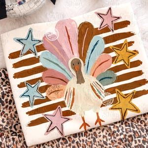 May include: A white t-shirt featuring a painted turkey graphic with a fanned tail of pink, blue, and cream feathers. The design includes horizontal brown brushstrokes and five stars in shades of pink, blue, and yellow surrounding the turkey.