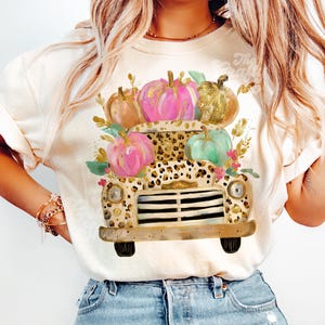 Pode incluir: Camiseta creme com uma cama de caminhão com estampa de leopardo cheia de abóboras coloridas. As abóboras são rosa, douradas e verdes, com detalhes florais. O caminhão tem um design vintage, perfeito para o outono.