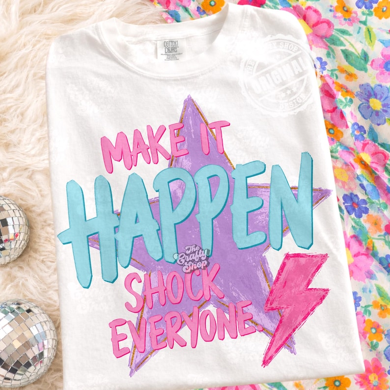 Może przedstawiać: Biała koszulka z napisem "MAKE IT HAPPEN SHOCK EVERYONE" w kolorze r&oacute;żowym i jasnoniebieskim. Projekt zawiera fioletową gwiazdę i r&oacute;żową błyskawicę. Koszulka ma okrągły dekolt i kr&oacute;tkie rękawy.