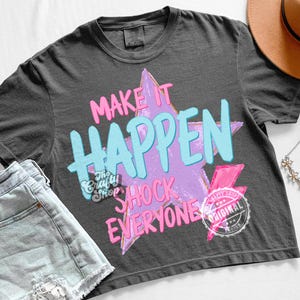 Może przedstawiać: Ciemnoszara koszulka z napisem "MAKE IT HAPPEN SHOCK EVERYONE" w r&oacute;żowych i niebieskich literach na fioletowym wzorze gwiazdy. Zawiera r&oacute;żową błyskawicę i okrągłe logo. Casualowy element mody.