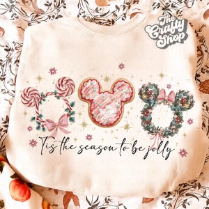 May include: Cream-colored sweatshirt with a festive design. Features three Mickey Mouse head wreaths: a candy cane wreath, a pink cookie, and a floral wreath. The text "Tis the season to be jolly" is written across the bottom.