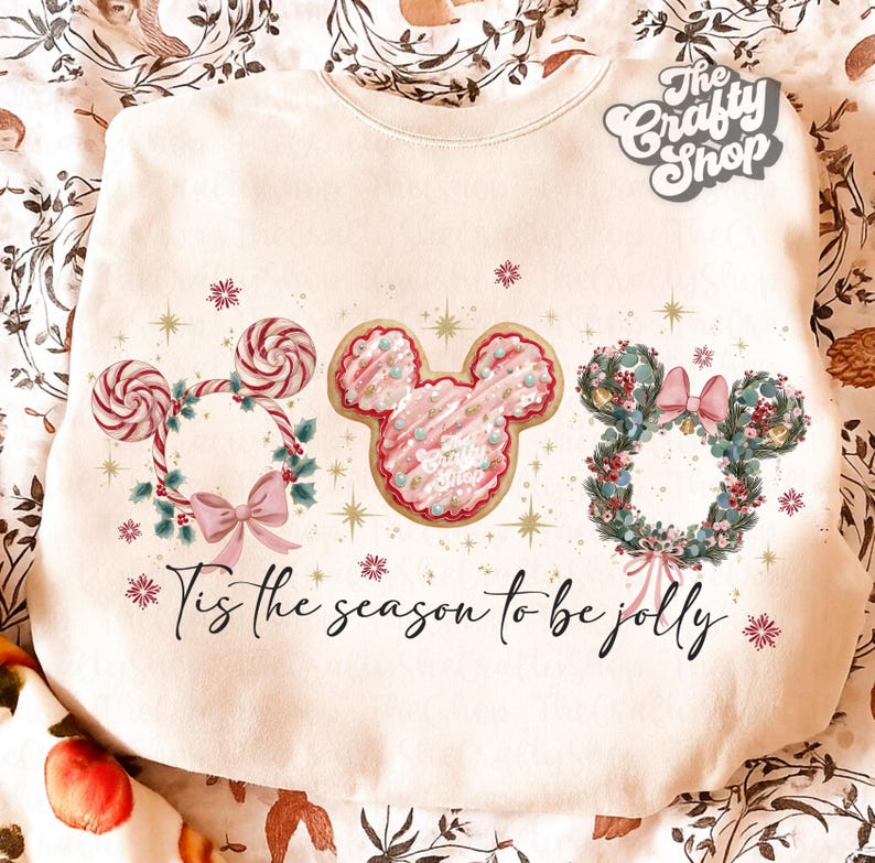 May include: Cream-colored sweatshirt with a festive design. Features three Mickey Mouse head wreaths: a candy cane wreath, a pink cookie, and a floral wreath. The text "Tis the season to be jolly" is written across the bottom.