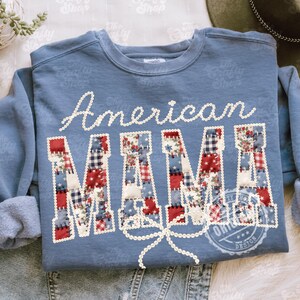 Puede incluir: Sudadera azul con la inscripci&oacute;n "American MAMA". La palabra "American" est&aacute; en escritura blanca, y "MAMA" est&aacute; en letras de patchwork con patrones rojos, blancos y azules. Una cinta blanca est&aacute; debajo de la palabra "MAMA".