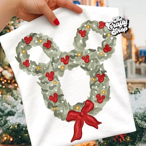 Pode incluir: Camiseta branca com um desenho de guirlanda de Natal em forma de Mickey Mouse. A guirlanda é composta por folhagens verdes, cabeças vermelhas do Mickey Mouse e pequenas luzes amarelas. Uma grande fita vermelha está na parte inferior. O texto "The Crafty Shop" está no canto superior direito.