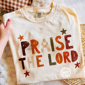 Peut inclure: Un t-shirt &agrave; manches courtes de couleur cr&egrave;me avec le texte "PRAISE THE LORD" imprim&eacute; dans une police en blocs multicolore. Le motif pr&eacute;sente diverses nuances de brun, d'orange et de vert, agr&eacute;ment&eacute; de petites &eacute;toiles. &Eacute;tiquette Comfort Colors.