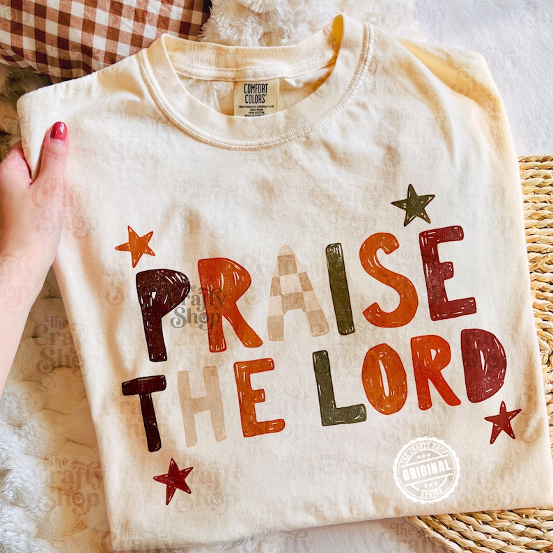 Peut inclure: Un t-shirt &agrave; manches courtes de couleur cr&egrave;me avec le texte "PRAISE THE LORD" imprim&eacute; dans une police en blocs multicolore. Le motif pr&eacute;sente diverses nuances de brun, d'orange et de vert, agr&eacute;ment&eacute; de petites &eacute;toiles. &Eacute;tiquette Comfort Colors.