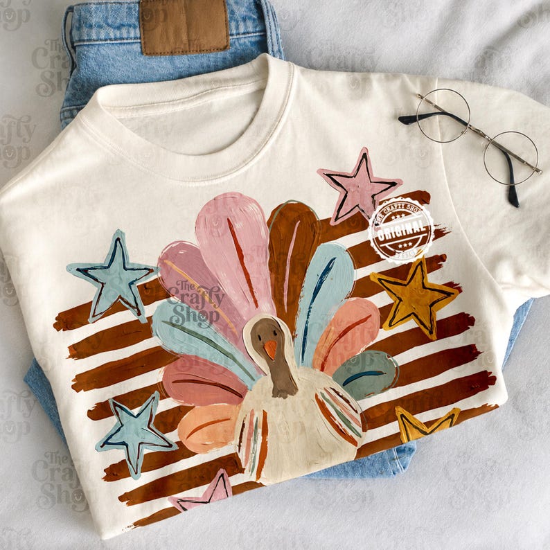 May include: A cream-coloured t-shirt featuring a graphic of a turkey with colourful, brushstroke-style feathers in shades of pink, blue, and brown. The design includes brown horizontal stripes, several five-pointed stars, and a circular logo.