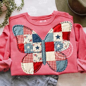 Pode incluir: Um moletom coral com um design de borboleta em patchwork. As asas da borboleta são compostas por quadrados de tecido vermelho, branco e azul, alguns com padrões de estrelas e flores. O desenho é costurado na camisa com linha azul.