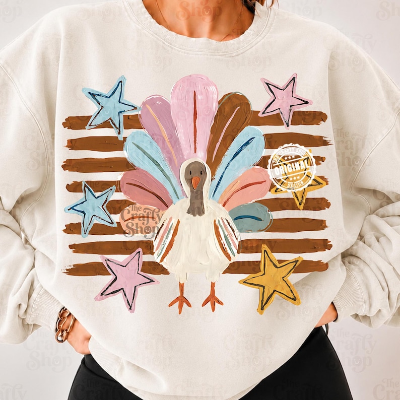 May include: A cream-coloured crewneck sweatshirt featuring a central graphic of a turkey with colourful, brushstroke-style tail feathers. The design includes horizontal brown brushstroke stripes and several five-pointed stars in blue, pink, and yellow.