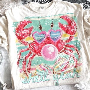 May include: Cream-colored t-shirt featuring a vibrant crab graphic. The crab is red and wears heart-shaped sunglasses. The shirt has the text "shell yeah" in a teal script, and the words "The Crafty Shop" and "Original" are also visible.