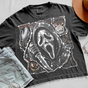 May include: A charcoal gray short-sleeve t-shirt featuring a graphic print of a masked figure holding a knife, surrounded by Halloween-themed imagery including pumpkins and corn. The design has a distressed, textured appearance with white highlights.