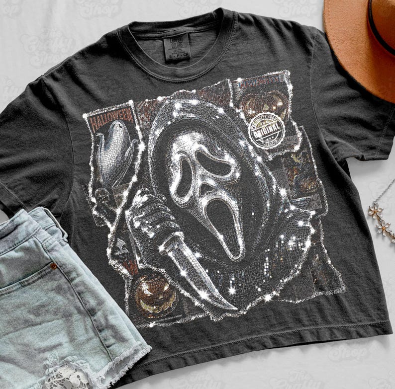 May include: A charcoal gray short-sleeve t-shirt featuring a graphic print of a masked figure holding a knife, surrounded by Halloween-themed imagery including pumpkins and corn. The design has a distressed, textured appearance with white highlights.