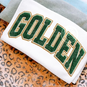 Pode incluir: Camisola branca com a palavra "GOLDEN" bordada em verde com contorno dourado. A camisola está dobrada, e o fundo inclui um padrão de leopardo e tecido de cor sólida.