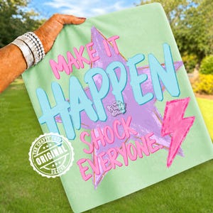 Może przedstawiać: Jasnozielona koszulka z napisami "MAKE IT HAPPEN SHOCK EVERYONE" w kolorze r&oacute;żowym i niebieskim. Projekt zawiera fioletową gwiazdę i r&oacute;żową błyskawicę. Widoczne jest r&oacute;wnież oryginalne logo The Crafty Shop.