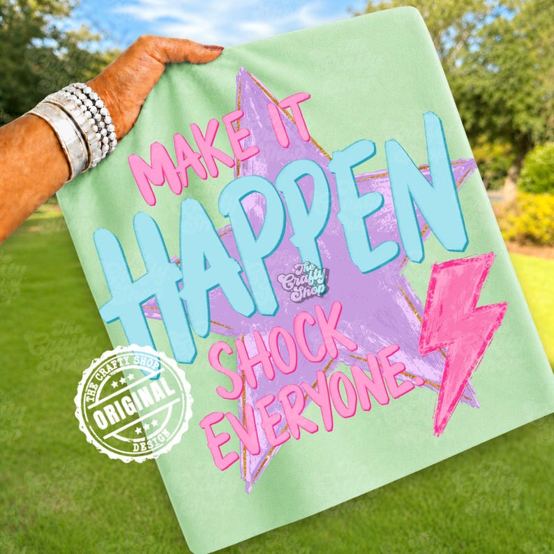 Może przedstawiać: Jasnozielona koszulka z napisami "MAKE IT HAPPEN SHOCK EVERYONE" w kolorze r&oacute;żowym i niebieskim. Projekt zawiera fioletową gwiazdę i r&oacute;żową błyskawicę. Widoczne jest r&oacute;wnież oryginalne logo The Crafty Shop.