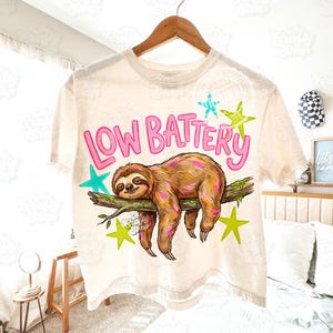 Puede incluir: Camiseta color crema con un gr&aacute;fico de un perezoso descansando en una rama, rodeado de estrellas. El texto "LOW BATTERY" es rosa. El perezoso es marr&oacute;n y beige con acentos rosas. La camiseta est&aacute; colgada en una percha de madera.