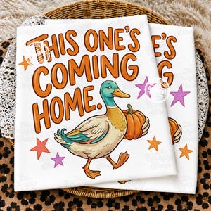 Pode incluir: Uma t-shirt branca com um gráfico de um pato-real segurando uma abóbora laranja. O texto diz THIS ONE'S COMING HOME em letras grandes cor de laranja. O design inclui pequenas estrelas laranja e roxas ao redor do pato e do texto.