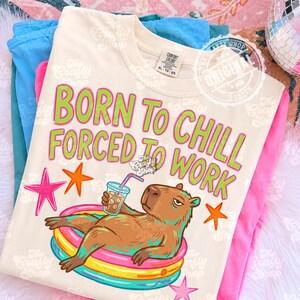 May include: Cream-colored t-shirt with the text "BORN TO CHILL FORCED TO WORK" in green and pink. The shirt features a cartoon capybara relaxing in an inflatable pool, holding a drink. Additional star graphics are included.