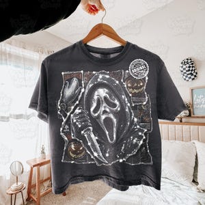 May include: A charcoal gray short-sleeve t-shirt featuring a graphic print of a hooded figure holding a knife, surrounded by Halloween-themed imagery including pumpkins and corn. A circular white label with the text Original is on the chest.