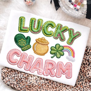 Pode incluir: Camiseta branca com design bordado "LUCKY CHARM". O design inclui uma luva verde com um trevo, um pote de ouro dourado, um trevo verde e um arco-íris. As letras são verdes e rosa.