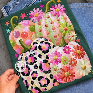 Pode incluir: Uma t-shirt verde-escura com uma impressão gráfica de três abóboras. Uma abóbora tem bolinhas cor-de-rosa e laranja, outra é verde-clara com pequenas abóboras cor de laranja e flores cor-de-rosa, e a terceira tem um padrão de leopardo rosa.