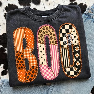 Pode incluir: Uma camisola de gola redonda cinzento-carvão com a palavra BOO em letras grandes estilo patchwork. Cada letra é composta por vários padrões, incluindo riscas cor de laranja, pintas, estampado de leopardo, xadrez vichy e flores.