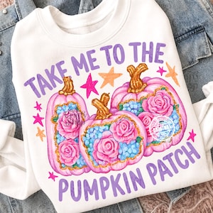 Pode incluir: Uma camisola branca com uma impressão gráfica de três abóboras cor-de-rosa decoradas com rosas cor-de-rosa e flores azuis. O texto "TAKE ME TO THE PUMPKIN PATCH" está impresso em roxo acima e abaixo, com estrelas cor de laranja e rosa.