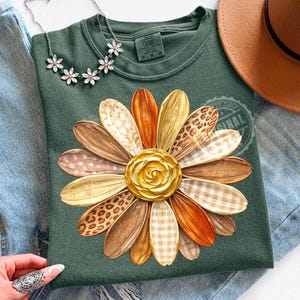 Pode incluir: Um moletom verde Comfort Colors com um grande design de margarida. As pétalas da margarida apresentam vários padrões e texturas em tons de castanho, laranja e bege, com um centro dourado. Um colar floral e um chapéu castanho também são visíveis.