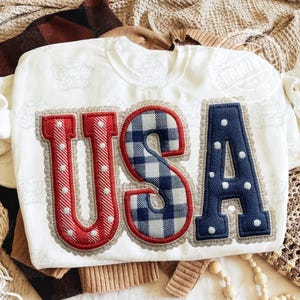 Pode incluir: Camisola branca com as letras "USA" aplicadas na frente. O "U" e o "A" são vermelhos com bolinhas brancas, e o "S" é azul e branco xadrez. As letras são contornadas com uma costura decorativa.