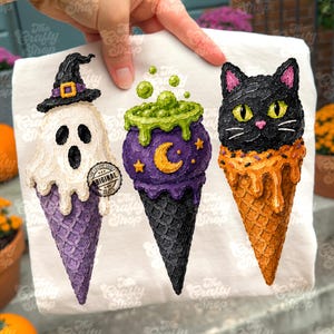 Pode incluir: Uma t-shirt branca com uma impressão gráfica de três cones de gelado. O primeiro é um fantasma branco com um chapéu de bruxa preto num cone roxo. O segundo é um caldeirão roxo com líquido verde a borbulhar. O terceiro é uma cabeça de gato preto num cone laranja.