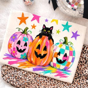 May include: A white rectangular item featuring a graphic of three jack-o'-lanterns and a black cat. The left pumpkin is pink with flowers, the center is orange with a black cat on top, and the right is a multicolored checkered pumpkin with stars.
