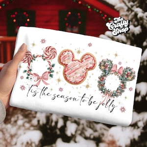May include: White holiday-themed towel featuring three Mickey Mouse-shaped designs: a candy cane wreath, a pink frosted cookie, and a floral wreath. The text "'Tis the season to be jolly" is written in cursive. The Crafty Shop logo is in the upper right corner.