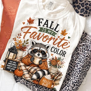 May include: A white t-shirt featuring a graphic of a raccoon wearing glasses and a sweater, holding a mug. The text reads FALL IS MY FAVORITE COLOR, COZY DAYS WARM DRINKS CRUNCHY LEAVES HAPPY HEART, and HELLO FALL. Includes autumn leaves, a pumpkin, and a lantern.