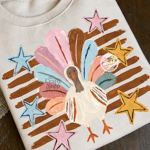 May include: A cream-coloured sweatshirt featuring a graphic print of a turkey with a fanned tail in shades of pink, blue, and brown. The design includes brown horizontal brushstrokes and five-pointed stars in pink, light blue, and yellow.