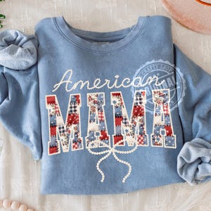 Puede incluir: Sudadera azul claro con las palabras "American MAMA" en letras blancas y letras de patchwork. Las letras est&aacute;n decoradas con patrones rojos, blancos y azules. Una cinta blanca est&aacute; debajo de la palabra "MAMA".