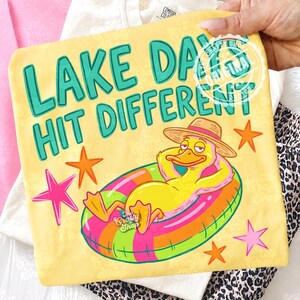 May include: Yellow t-shirt with the text "LAKE DAYS HIT DIFFERENT" in teal. The design features a cartoon duck in a straw hat, relaxing on a colourful inflatable ring. Pink and orange stars are scattered around the duck.