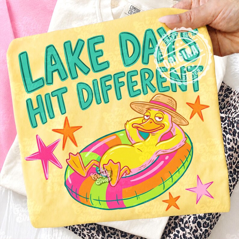 May include: Yellow t-shirt with the text "LAKE DAYS HIT DIFFERENT" in teal. The design features a cartoon duck in a straw hat, relaxing on a colourful inflatable ring. Pink and orange stars are scattered around the duck.