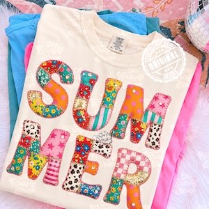 Pode incluir: Camiseta creme com a palavra "SUMMER" em letras de patchwork coloridas. As letras apresentam padrões florais, de bolinhas e listrados. A camiseta está dobrada com outras camisetas coloridas.